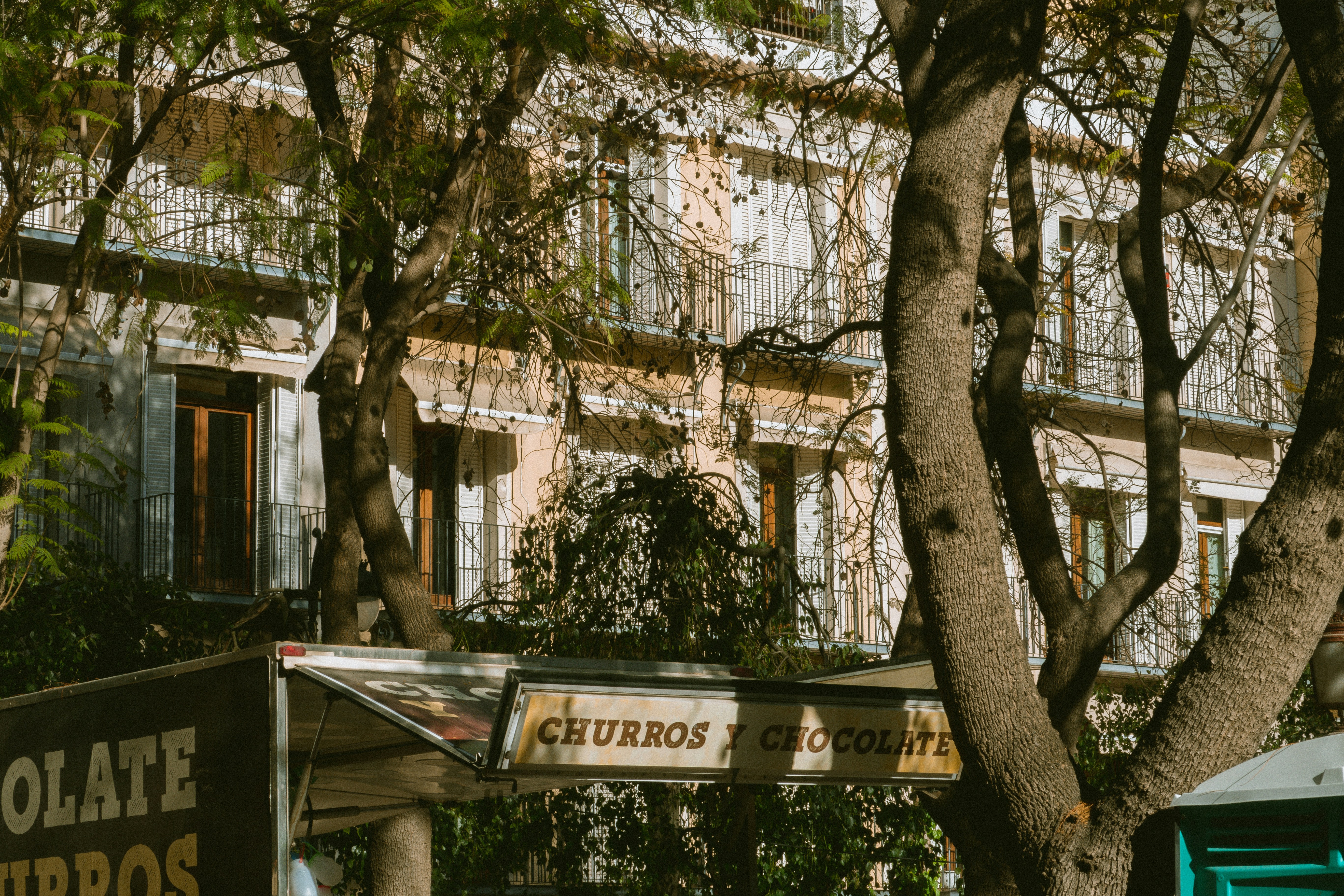 A churros and chocolate sign on a shaded street, evoking Spain&rsquo;s caf&eacute; and sweet-snack culture.